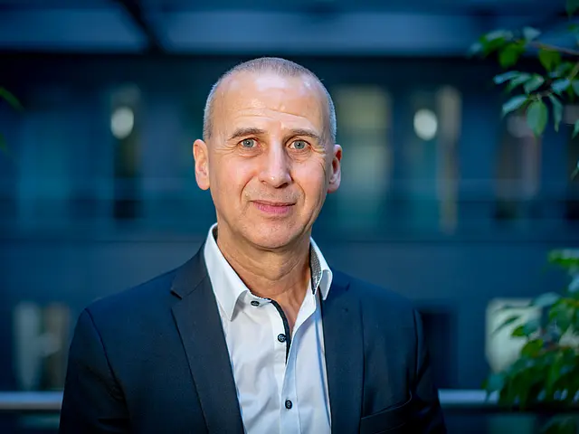 Andrej Heilmann in Business‑Kleidung steht vor einem modernen Gebäude, umgeben von Pflanzen im Vordergrund.