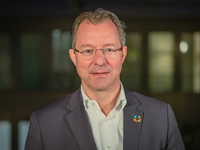 Oliver Zernahle in dunklem Anzug und weißem Hemd steht vor einem Gebäude; am Revers befindet sich ein farbiges Pin‑Abzeichen.