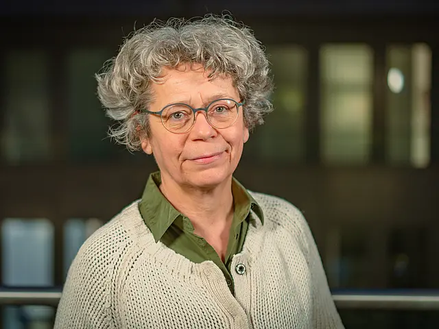Annette Siering mit grauem, lockigem Haar, grünem Hemd und hellem Strickpullover steht vor einem modernen Gebäude mit beleuchteten Fenstern.
