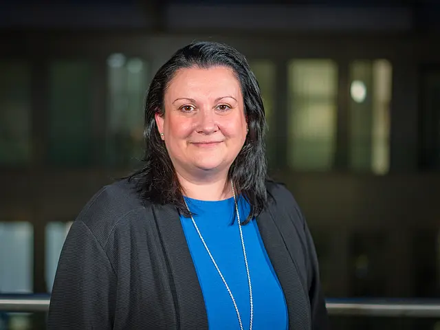 Sabrina Poweleit in blauem Oberteil und dunkler Jacke steht vor einem modernen Gebäude mit beleuchteten Fenstern im Hintergrund.