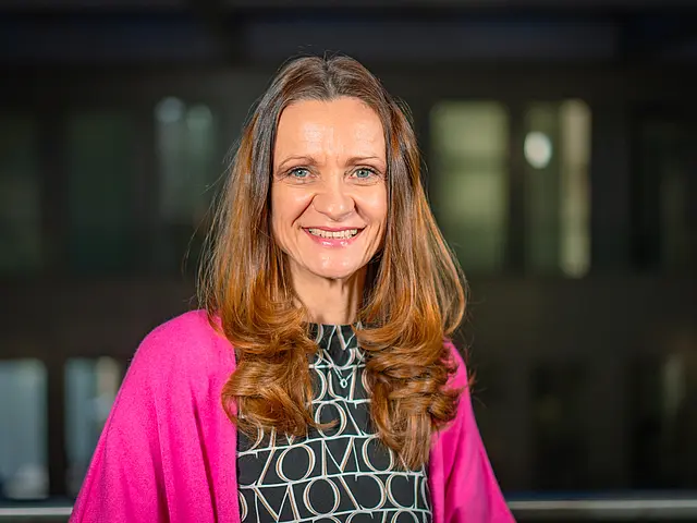 Claudia Rathfux mit langen Haaren, gemustertem Oberteil und pinkem Schal steht vor einem modernen Gebäude mit beleuchteten Fenstern.